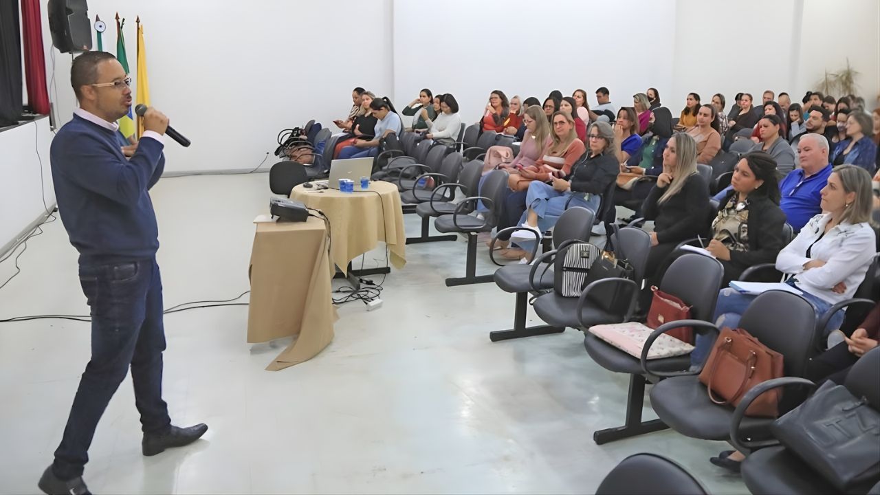 Imagem mostra o professor Julio Passos com um microfone na mão ministrando uma palestra sobre inteligencia artificial para outros professores em um auditório lotado.
